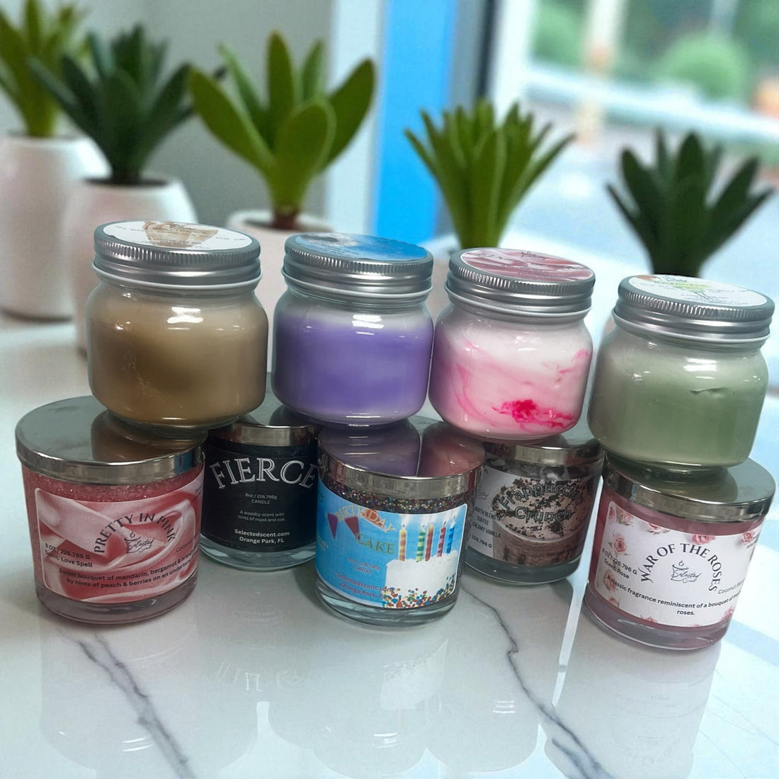 Collection of long lasting natural candles in various colors and scents displayed on a table with green plants in the background