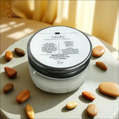 Selected Scent Cheers Massage Candle with Mango Butter in a clear jar with silver lid surrounded by smooth brown stones